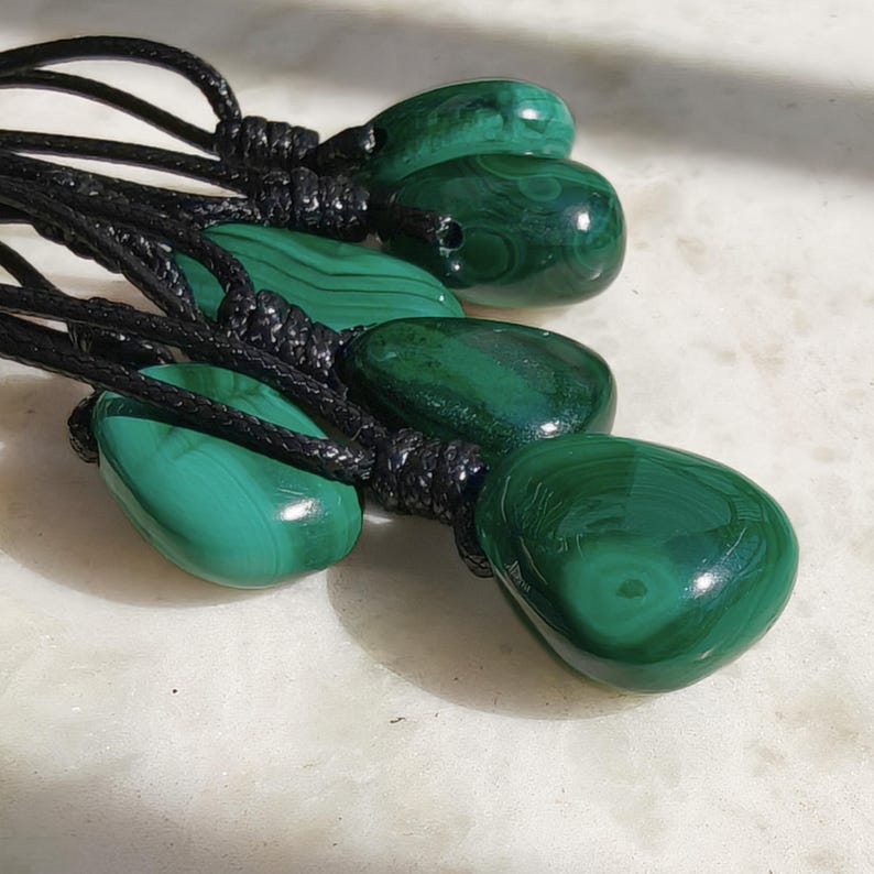 Close-up of multiple polished irregular Malachite in shades of vivid green and deep forest green, arranged on a light stone surface. The green opaque gemstones have smooth glossy surfaces with distinct banded patterns, concentric lines, and varied formations, including rounded and triangular shapes with layered green stripes and darker swirls. It is attached to a black cord with a top knot and adjustable knot closure. Malachite is associated with transformation, protection, and emotional healing