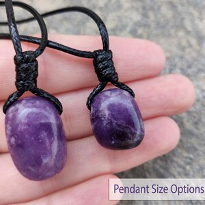 May include: Two amethyst pendant necklaces. Each necklace features a polished, oval-shaped purple gemstone suspended from a black cord. The cords are knotted at the top. The image includes the text "Pendant Size Options".