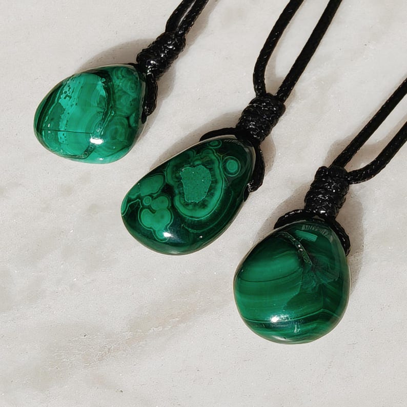 Close-up of 3 polished irregular Malachite in shades of vivid green and deep forest green, arranged on a light stone surface. The green opaque gemstones have smooth glossy surfaces with distinct banded patterns, concentric lines, and varied formations, including rounded and triangular shapes with layered green stripes and darker swirls. It is attached to a black cord with a top knot and adjustable knot closure. Malachite is associated with transformation, protection, and emotional healing.