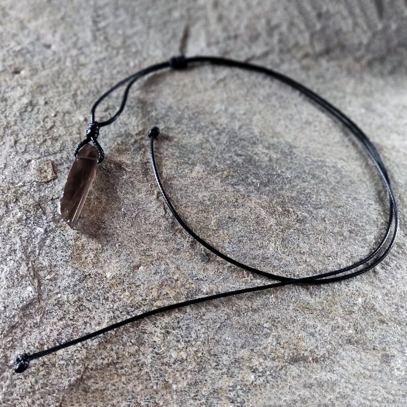 One raw smoky quartz necklace with rectangular stone in dark brown translucent hues, rough-textured with irregular edges, hung on a knotted black cord, placed on a light brown stone surface with a natural grainy texture. Smokey Quartz is associated with grounding, protection, and emotional calmness.