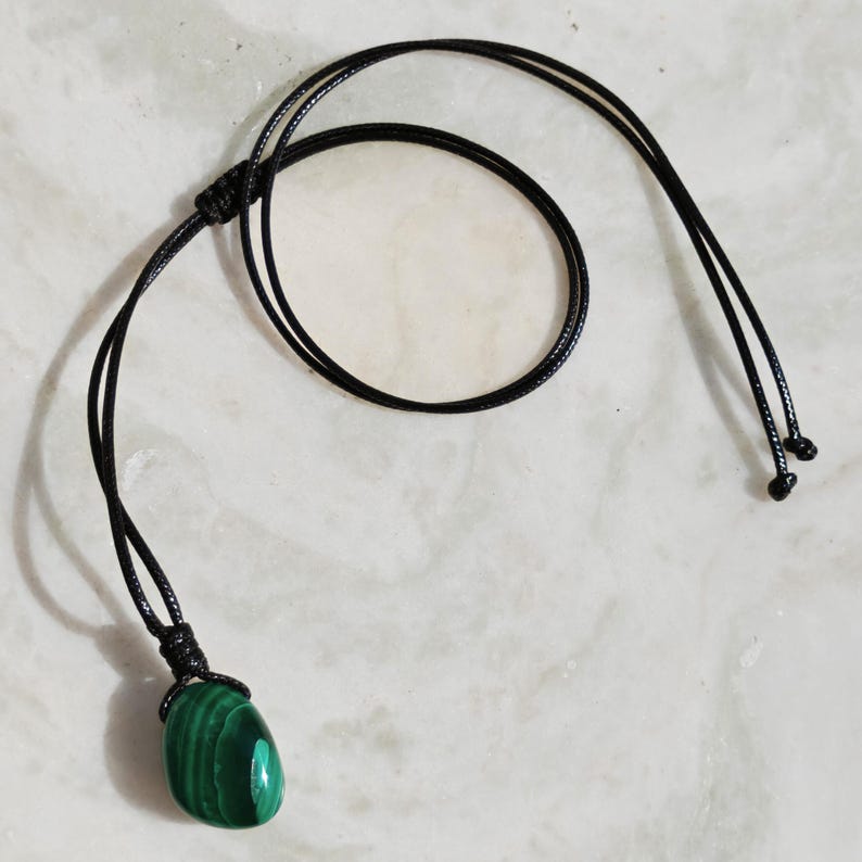 Close-up of a polished oval Malachite in shades of vivid green and deep forest green, resting on a light stone surface. The green opaque gemstone has a smooth glossy surface with distinct concentric banding, circular eye-like formations, and layered green gradients, with light reflections highlighting its rounded shape. It is attached to a black cord with a top knot and adjustable knot closure. Malachite is associated with transformation, protection, and emotional healing.