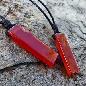 Two polished, elongated, rectangular Carnelian crystal necklaces are displayed on a textured gray surface. The orange-red gemstones have a smooth, translucent texture. There are multiple pendants, each attached to a black cord. Carnelian is associated with courage, vitality, and motivation.