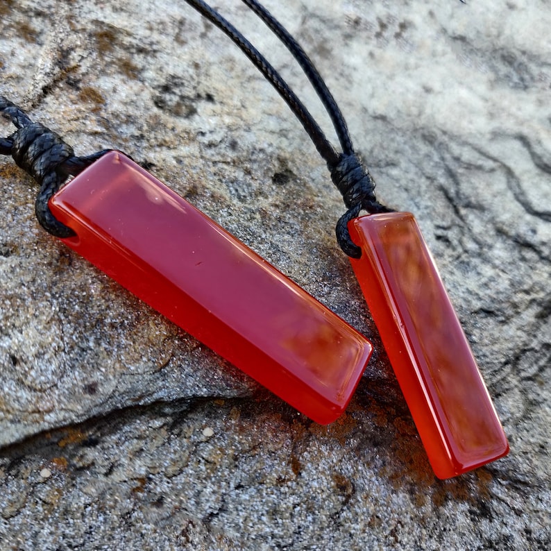 Two polished, elongated, rectangular Carnelian crystal necklaces are displayed on a textured gray surface. The orange-red gemstones have a smooth, translucent texture. There are multiple pendants, each attached to a black cord. Carnelian is associated with courage, vitality, and motivation.