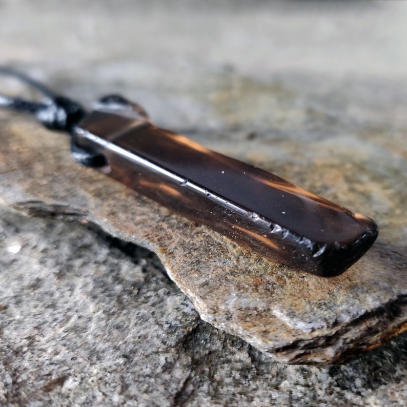 One raw smoky quartz necklace with rectangular stone in dark brown translucent hues, rough-textured with irregular edges, hung on a knotted black cord, placed on a light brown stone surface with a natural grainy texture. Smokey Quartz is associated with grounding, protection, and emotional calmness.