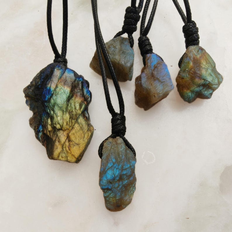 Close-up of 5 raw irregular Labradorite stones in shades of blue, teal, green, and golden hues, displayed on a light surface. The Labradorite gemstones have a rough natural texture with strong iridescent flashes, layered color play, and visible striations across their surfaces. Each stone is secured with a top knot and attached to a black cord with adjustable knot closure. Labradorite is associated with transformation, protection, intuition, and spiritual shielding.