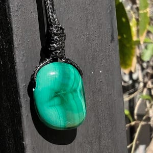 A polished irregular Malachite in shades of green and light emerald green, hanging against a dark textured surface with blurred greenery in the background. The green opaque gemstone has a smooth glossy surface with soft banded patterns, subtle linear striations, and gentle color gradients, with light reflections highlighting its rounded form. It is attached to a black cord with a top knot and adjustable knot closure. Malachite is associated with transformation, protection, and emotional healing.