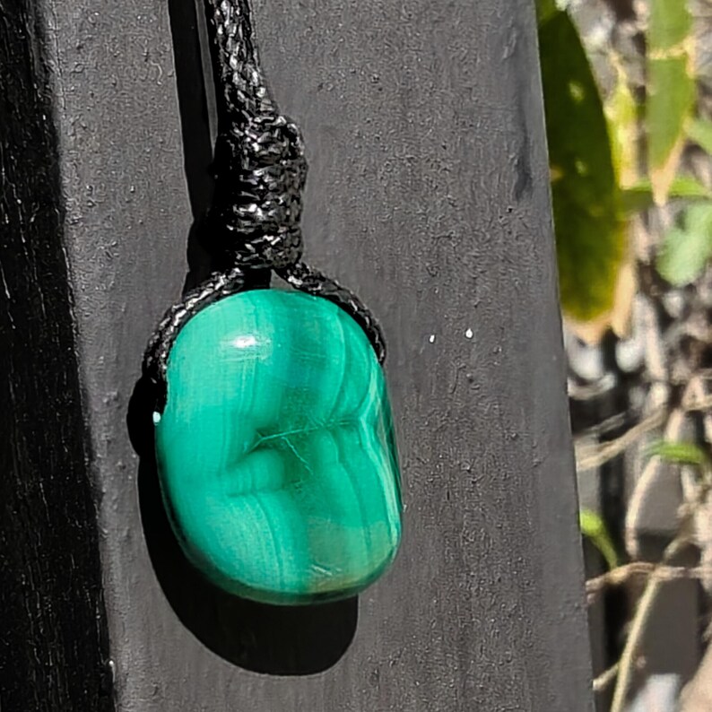 A polished irregular Malachite in shades of green and light emerald green, hanging against a dark textured surface with blurred greenery in the background. The green opaque gemstone has a smooth glossy surface with soft banded patterns, subtle linear striations, and gentle color gradients, with light reflections highlighting its rounded form. It is attached to a black cord with a top knot and adjustable knot closure. Malachite is associated with transformation, protection, and emotional healing.