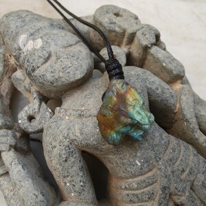 Close-up of a raw irregular Labradorite in shades of blue, teal, green, and golden brown, displayed on a carved stone statue. The Labradorite gemstone has a natural rough texture with strong iridescent flashes, visible striations, and layered color play with reflective highlights. It is secured with a top knot and attached to a black cord with adjustable knot closure. Labradorite is associated with transformation, protection, intuition, and inner strength.