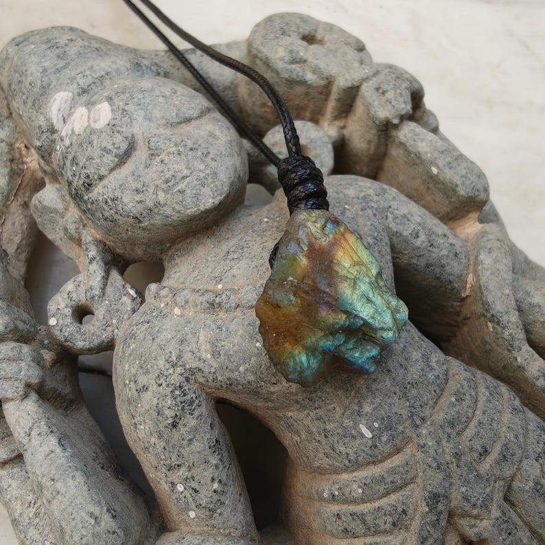 Close-up of a raw irregular Labradorite in shades of blue, teal, green, and golden brown, displayed on a carved stone statue. The Labradorite gemstone has a natural rough texture with strong iridescent flashes, visible striations, and layered color play with reflective highlights. It is secured with a top knot and attached to a black cord with adjustable knot closure. Labradorite is associated with transformation, protection, intuition, and inner strength.