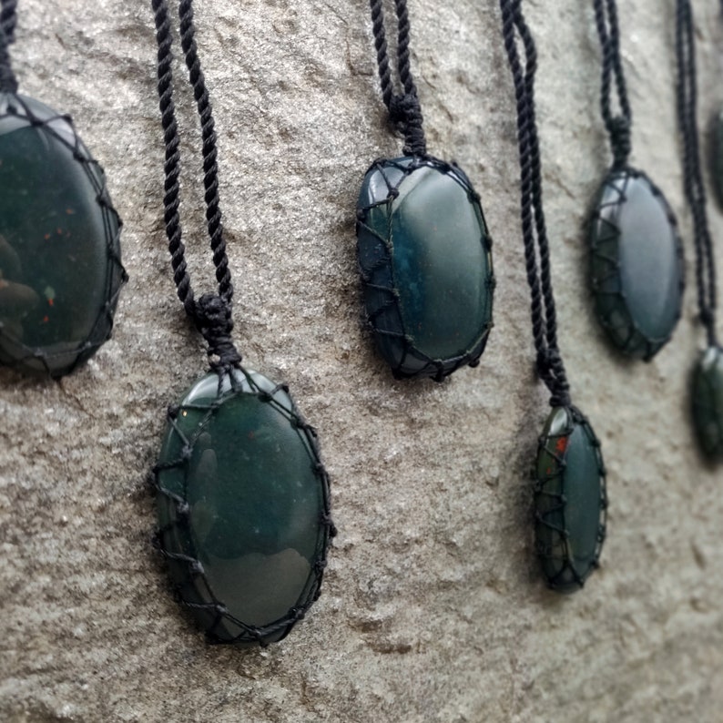 Several Bloodstone pendant necklaces displayed hanging from adjustable twisted black cords with a sliding knot closure against a textured natural stone wall. Each polished oval gemstone features deep forest-green tones with subtle natural red mineral inclusions, making every pendant unique. Bloodstone is associated with Courage, Resilience, Grounding, Vitality, and Inner Strength.
