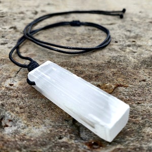 One raw, rectangular selenite pendant hang from black cord necklaces, rest on a textured gray stone surface background. The translucent white crystal have a fibrous, slightly uneven texture and catch the light. These selenite pendants are associated with clarity and purification.