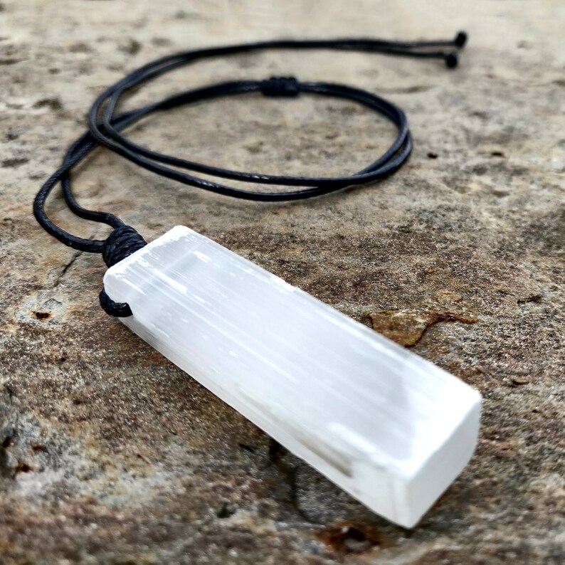 One raw, rectangular selenite pendant hang from black cord necklaces, rest on a textured gray stone surface background. The translucent white crystal have a fibrous, slightly uneven texture and catch the light. These selenite pendants are associated with clarity and purification.
