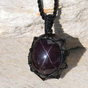 Close-up of a polished star garnet pendant in deep burgundy, wine-red, and dark plum tones, displayed on a light stone background. A distinct four-rayed star appears across the gemstone when illuminated, creating a bright reflective cross. The garnet has a smooth glossy finish with rich color depth and natural translucency. It is secured in a black macrame net and attached to a black adjustable cord. Associated with passion, vitality, grounding, courage, and emotional strength.