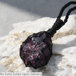 A single, raw, drop shaped raw Garnet necklace pendant is displayed on a textured white rock surface. The dark reddish-purple gemstone has a rough, unpolished texture. It is encased in a black macrame net that forms the necklace cord. Garnet is associated with passion, energy, protection, and healing.