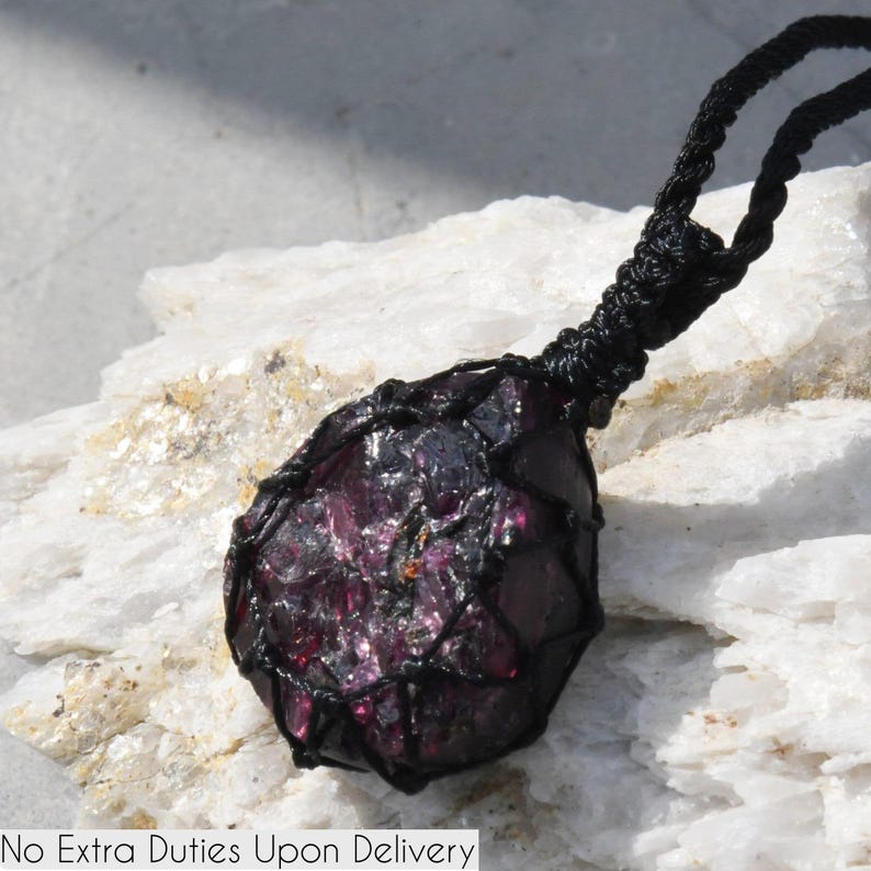 A single, raw, drop shaped raw Garnet necklace pendant is displayed on a textured white rock surface. The dark reddish-purple gemstone has a rough, unpolished texture. It is encased in a black macrame net that forms the necklace cord. Garnet is associated with passion, energy, protection, and healing.
