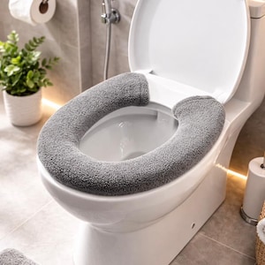 May include: A white toilet with a gray, plush toilet seat cover. The toilet seat is up, revealing the bowl. The bathroom has gray tile, a small plant, and a roll of toilet paper.