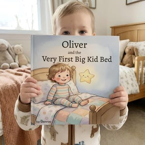 May include: A child holds a book titled "Oliver and the Very First Big Kid Bed." The book cover features an illustration of a child in pajamas on a bed with a glowing star. The background includes a bed and stuffed animals.