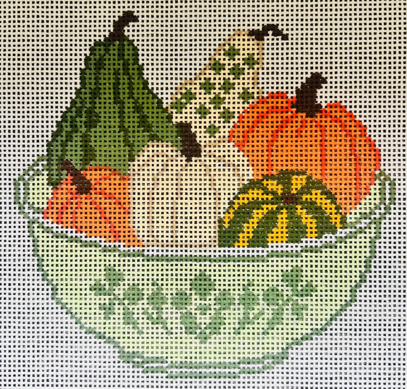 May include: A needlepoint canvas featuring a light green bowl with a delicate floral pattern, filled with a variety of colorful gourds and pumpkins in shades of orange, dark green, and cream, all set against a white grid background.