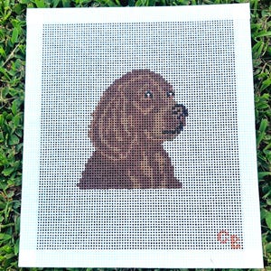 May include: A square needlepoint canvas featuring a brown dog profile design centered on a white grid. The dog has dark brown fur and a black nose. The letters CB are stitched in red in the bottom right corner of the white mesh fabric.
