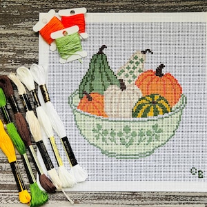 May include: A needlepoint canvas featuring a cross-stitch design of various gourds and pumpkins in a patterned bowl, accompanied by several skeins of embroidery floss in shades of yellow, green, brown, white, orange, and light green.