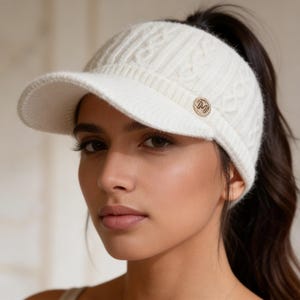 May include: Cream-colored knit visor with a cable knit pattern and a small, round gold-tone emblem. The visor has a curved brim and is designed to be worn with a ponytail. A stylish accessory for sun protection.