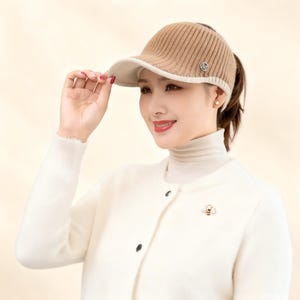 May include: A tan and cream-coloured visor hat with a ribbed texture. The hat has a small metal emblem on the side. The woman in the image is wearing a white turtleneck and cardigan.