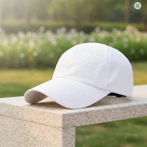 May include: A white baseball cap with a curved brim. The cap has a perforated design for breathability. The cap is sitting on a stone surface. The background is blurred with green foliage.