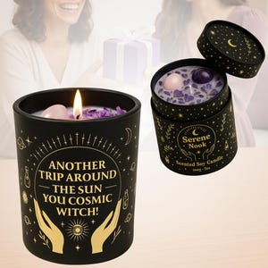 May include: Black soy candle with the text "ANOTHER TRIP AROUND THE SUN YOU COSMIC WITCH!" and a lit wick. The candle is decorated with gold celestial designs. A second candle in a box is also visible, labeled "Serene Nook".