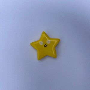 K&ouml;nnte beinhalten: Ein leuchtend gelbes, sternf&ouml;rmiges Objekt mit einer glatten, gl&auml;nzenden Oberfl&auml;che. Der Stern hat ein einfaches, handgezeichnetes Gesicht, bestehend aus zwei wei&szlig;en, kreisf&ouml;rmigen Augen mit schwarzen Pupillen und einem kleinen schwarzen Mund.