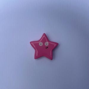 K&ouml;nnte beinhalten: Ein gl&auml;nzendes, f&uuml;nfzackiges sternf&ouml;rmiges Objekt in einem leuchtenden Pinkton. Der Stern zeigt ein Gesicht im Cartoon-Stil mit zwei wei&szlig;en, kreisf&ouml;rmigen Augen mit schwarzen Pupillen und einem kleinen, einfachen schwarzen Mundbogen.