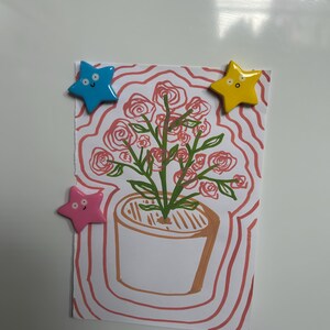 K&ouml;nnte beinhalten: Ein rechteckiges wei&szlig;es Papier mit einer handgezeichneten Illustration einer Topfpflanze mit rosa Rosen und gr&uuml;nen Stielen. Die Zeichnung ist von wellenf&ouml;rmigen rosa Linien umgeben und wird von drei sternf&ouml;rmigen Magneten in Blau, Gelb und Rosa gehalten.