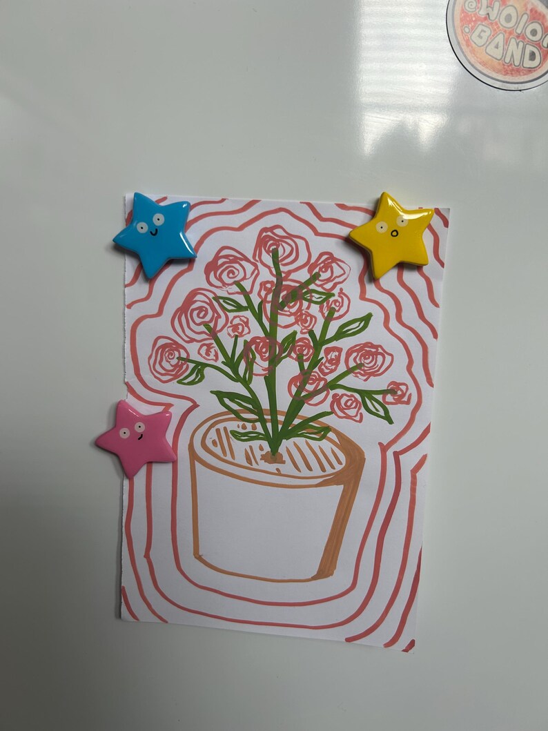 K&ouml;nnte beinhalten: Ein rechteckiges wei&szlig;es Papier mit einer handgezeichneten Illustration einer Topfpflanze mit rosa Rosen und gr&uuml;nen Stielen. Die Zeichnung ist von wellenf&ouml;rmigen rosa Linien umgeben und wird von drei sternf&ouml;rmigen Magneten in Blau, Gelb und Rosa gehalten.