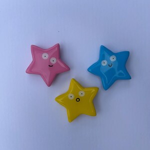 K&ouml;nnte beinhalten: Drei sternf&ouml;rmige Ornamente mit gl&auml;nzender Oberfl&auml;che in Pink, Blau und Gelb. Jeder Stern hat ein einzigartiges, handgemaltes Gesicht mit zwei Augen und einem Mund. Der pinke Stern schaut zur Seite, der blaue Stern l&auml;chelt und der gelbe Stern hat einen &uuml;berraschten Ausdruck mit offenem Mund.