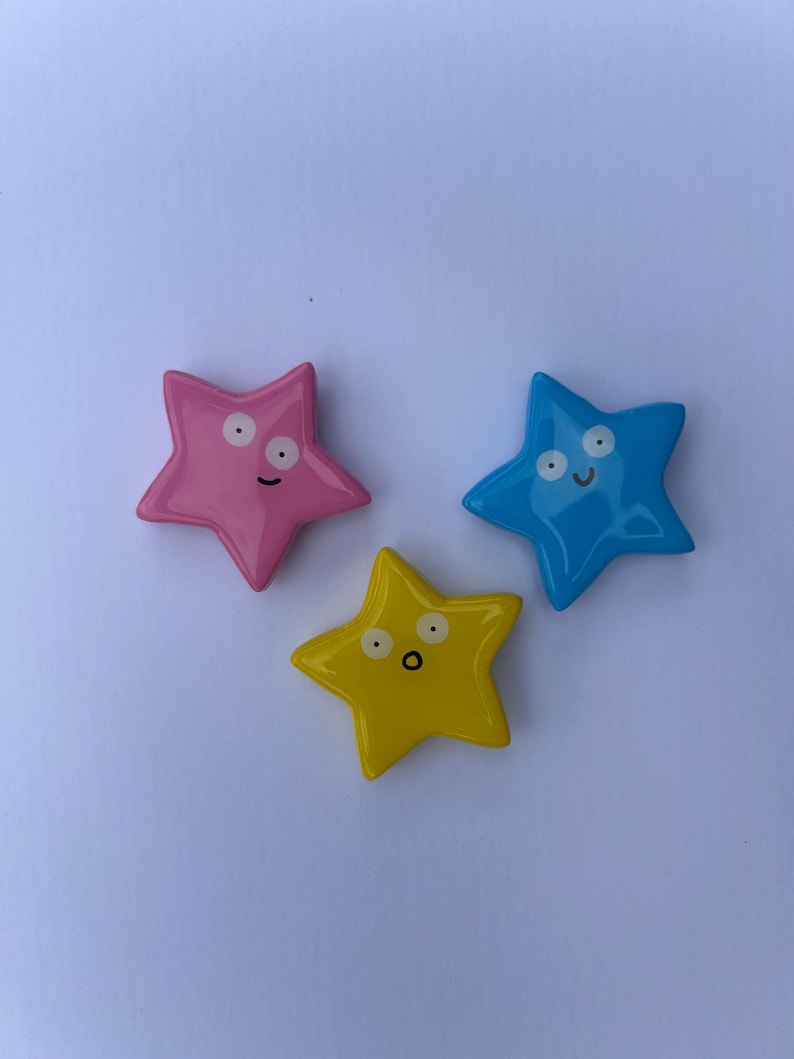 K&ouml;nnte beinhalten: Drei sternf&ouml;rmige Ornamente mit gl&auml;nzender Oberfl&auml;che in Pink, Blau und Gelb. Jeder Stern hat ein einzigartiges, handgemaltes Gesicht mit zwei Augen und einem Mund. Der pinke Stern schaut zur Seite, der blaue Stern l&auml;chelt und der gelbe Stern hat einen &uuml;berraschten Ausdruck mit offenem Mund.