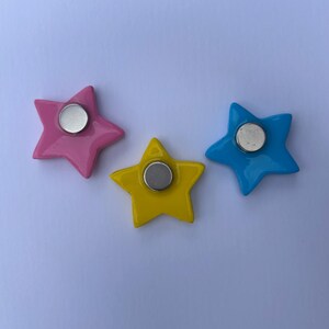 K&ouml;nnte beinhalten: Drei sternf&ouml;rmige Magnete in Pink, Gelb und Blau. Jeder Stern hat eine glatte, gl&auml;nzende Oberfl&auml;che und einen kreisf&ouml;rmigen, silberfarbenen Metallmagneten, der in der Mitte der R&uuml;ckseite befestigt ist. Die Magnete liegen locker auf einer Fl&auml;che.