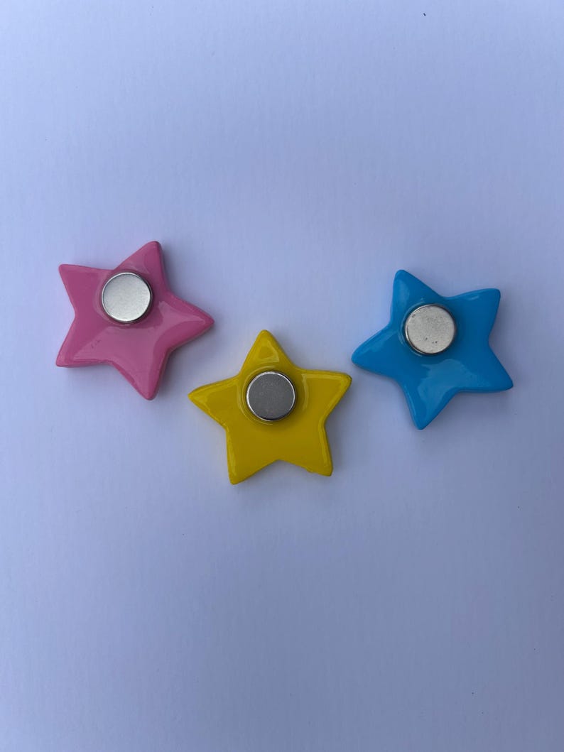 K&ouml;nnte beinhalten: Drei sternf&ouml;rmige Magnete in Pink, Gelb und Blau. Jeder Stern hat eine glatte, gl&auml;nzende Oberfl&auml;che und einen kreisf&ouml;rmigen, silberfarbenen Metallmagneten, der in der Mitte der R&uuml;ckseite befestigt ist. Die Magnete liegen locker auf einer Fl&auml;che.