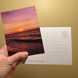 May include: A postcard featuring a vibrant sunset over the ocean, with shades of orange, pink, and purple in the sky. The text "Sunrise over Cape Cod Bay" is printed at the bottom. The back of the postcard has lines for writing and a space for a stamp.