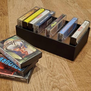Puede incluir: Un soporte negro para cintas de cassette lleno de varias cintas. Las cintas están en estuches de plástico transparente, con carátulas visibles. Algunas cintas están apiladas frente al soporte. Las carátulas incluyen nombres de bandas y álbumes.