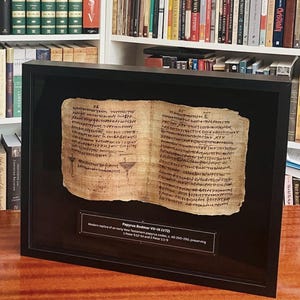 May include: Framed replica of the Papyrus Bodmer VII-IX (P72), an early New Testament papyrus codex. The ancient text is displayed in a black frame, with a descriptive plaque below. The text is in a faded brown color.