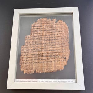 May include: Framed replica of a fragment from the Book of Genesis (R962). The text is written on a tan papyrus-like material and is enclosed in a white frame. The text at the bottom reads: "Modern Replica of the fragment from a folio of the Book of Genesis (R962) Produced in Egypt and dated to the 3rd".