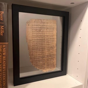 May include: A rectangular black frame containing a piece of light brown papyrus with dark ink Greek text. The papyrus has uneven, frayed edges and is displayed between two clear glass panes, showing the handwritten script clearly in the center.
