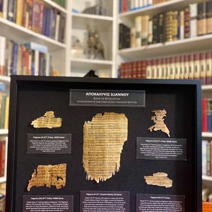 Puede incluir: Una colección enmarcada de réplicas de fragmentos de manuscritos antiguos del Libro de la Revelación. La exhibición incluye varios fragmentos de papiro con texto, etiquetados con detalles como "Papiro 24" y "Papiro 47". El marco es negro.