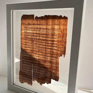 May include: A framed ancient papyrus fragment. The papyrus is a warm brown color with visible text in dark ink. The frame is a simple, light gray color, and the papyrus is protected by clear glass.