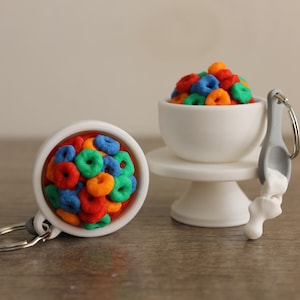May include: Miniature cereal keychains. One keychain features a white bowl filled with colorful cereal rings. Another keychain shows a white teacup on a pedestal, also filled with cereal. A gray spoon with a white substance is attached to the teacup.