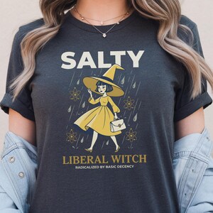 May include: A dark gray t-shirt featuring a graphic of a witch in a yellow dress and hat holding a handbag. The text reads SALTY LIBERAL WITCH and RADICALIZED BY BASIC DECENCY. The design includes rain drops and atom symbols in a light yellow color.