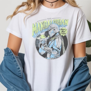 May include: White t-shirt featuring a circular graphic of The Mandalorian holding Grogu, with text that reads "THE MANDALORIAN" and "THAT'S NOT A TOY!". The design is in shades of blue, green, and gray.