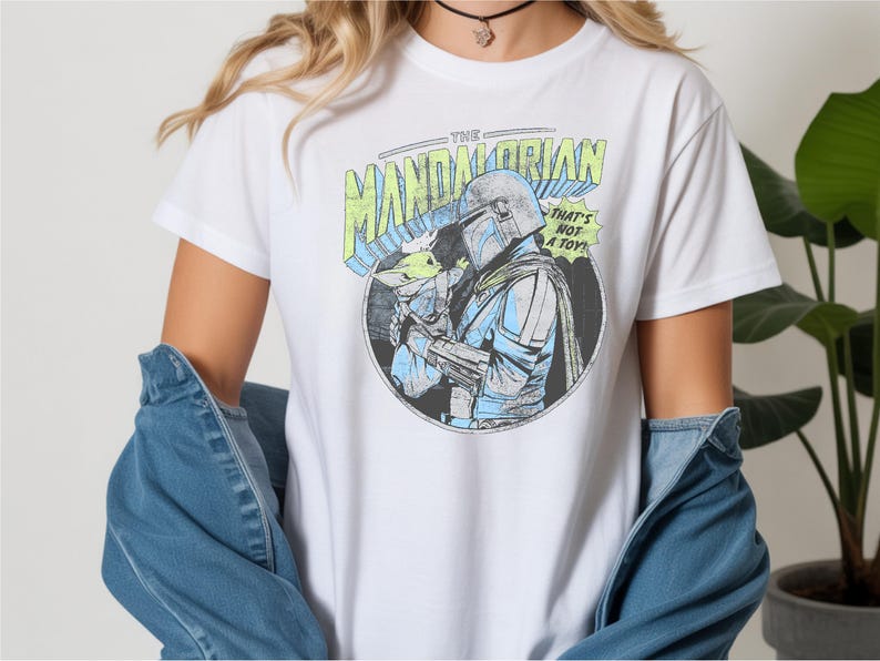 May include: White t-shirt featuring a circular graphic of The Mandalorian holding Grogu, with text that reads "THE MANDALORIAN" and "THAT'S NOT A TOY!". The design is in shades of blue, green, and gray.