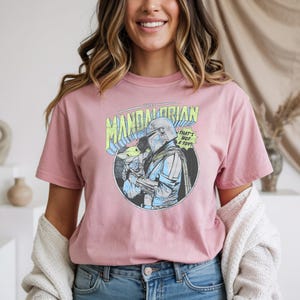 May include: A dusty rose t-shirt featuring a circular graphic of The Mandalorian holding Grogu, with the text "THE MANDALORIAN" and "THAT'S NOT A TOY!". The graphic is in blue, green, and yellow. The shirt is made of soft material.