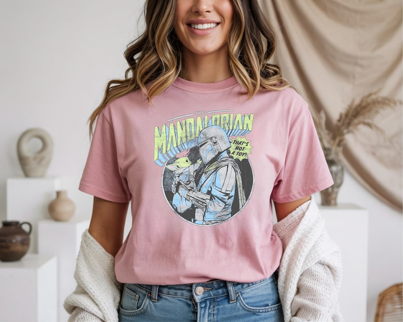 May include: A dusty rose t-shirt featuring a circular graphic of The Mandalorian holding Grogu, with the text "THE MANDALORIAN" and "THAT'S NOT A TOY!". The graphic is in blue, green, and yellow. The shirt is made of soft material.