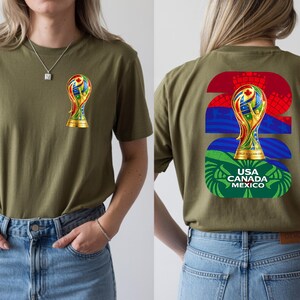 May include: Olive green t-shirt featuring a colorful trophy graphic on the front and back. The back design includes the number "22" in red and blue, the trophy, and the text "USA CANADA MEXICO".